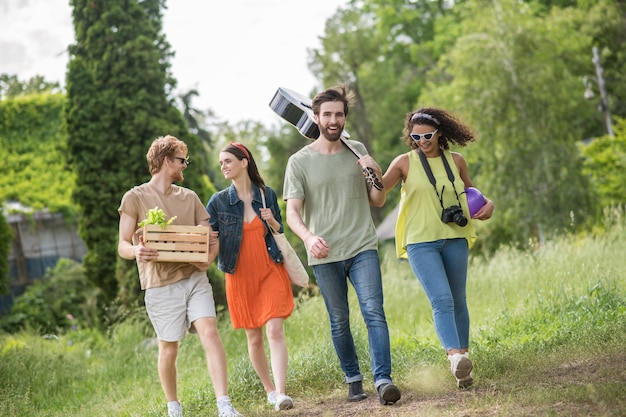 Moment heureux. Jeunes gars heureux avec de jolies filles bras dessus bras dessous marchant sur un pique-nique dans un parc verdoyant par une chaude journée ensoleillée