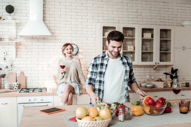 Moment joyeux. Séduisante jeune homme aux cheveux noirs et son sourire aux cheveux longs tout en buvant du vin rouge