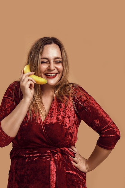 Moment ludique tourné en studio d'une femme souriante de grande taille en robe de velours rouge tenant une banane