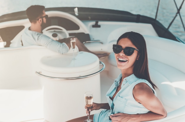 Moment parfait ensemble. Belle jeune femme tenant un verre de champagne et regardant la caméra pendant que son petit ami conduit un yacht