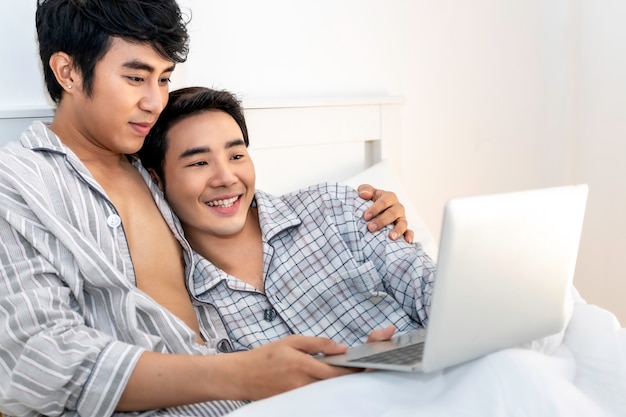 Moment romantique Couple asiatique homosexuel en pyjama shopping en ligne avec ordinateur portable dans le lit