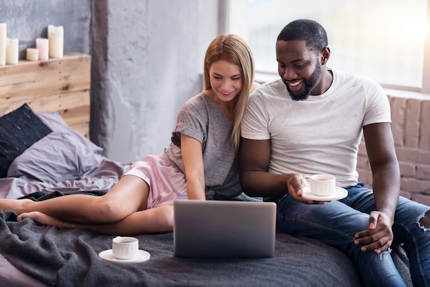 Moments joyeux. Doux jeune couple international couché dans la chambre tout en utilisant un ordinateur portable et en buvant du thé.