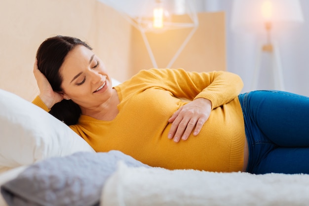 Mon bébé. Heureuse brune souriante tout en regardant avec impatience et allongée sur le lit dans sa chambre