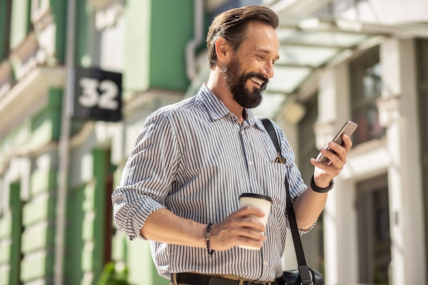 Sur mon chemin pour aller au travail. Tour de taille d'un homme barbu joyeux à l'aide de son smartphone et de boire du café