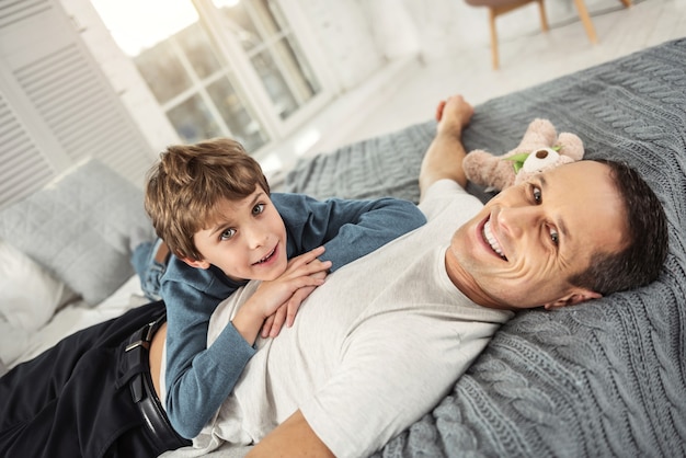 Mon cher papa. Beau garçon blond joyeux souriant et son papa allongé sur le dos sur le canapé et ils se détendent ensemble