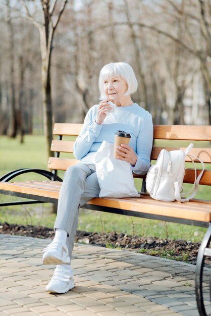 Mon déjeuner. Alerte vieille femme appréciant son café et un cookie assis sur le banc