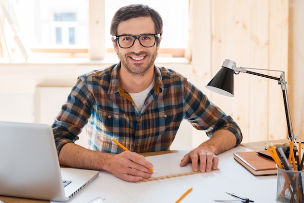 Mon espace pour créer. Beau jeune homme concevant un projet et souriant à la caméra alors qu'il était assis sur son lieu de travail