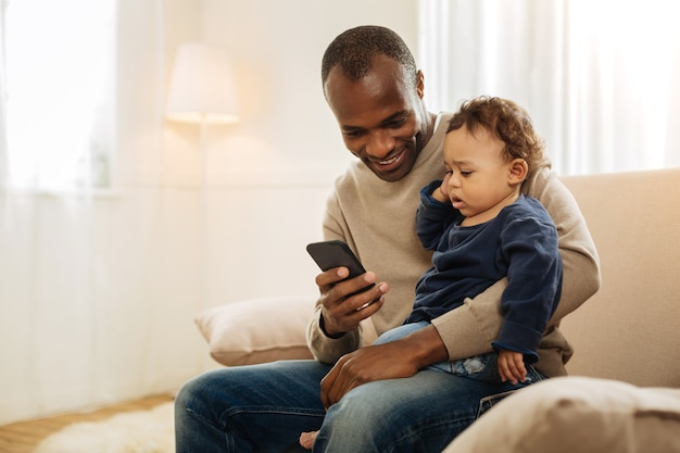 Mon fils. Alerte homme afro-américain aux cheveux noirs souriant montrant et quelque chose au téléphone à son fils tandis que l'enfant assis sur ses genoux