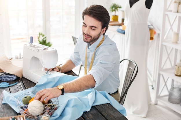 Mon inspiration. Ravi tailleur aux cheveux noirs souriant et travaillant sur une machine à coudre