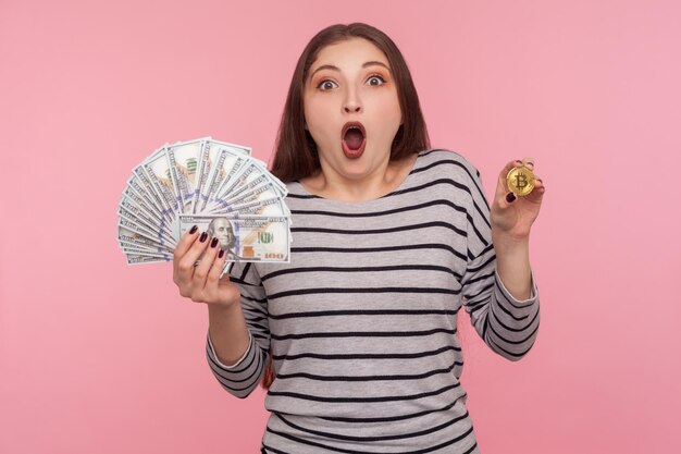 Monnaie électronique, crypto-monnaie, Portrait d'une femme étonnée en sweat-shirt rayé tenant des billets d'un dollar et du bitcoin, regardant la caméra avec une expression choquée. tourné en studio isolé sur fond rose