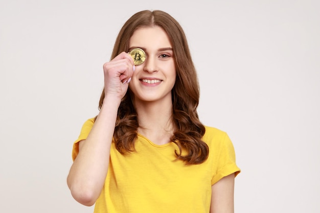 Monnaie numérique, crypto-monnaie. Jolie femme de jeune âge en T-shirt jaune de style décontracté, couvrant les yeux avec du bitcoin doré, regardant la caméra. Tourné en studio intérieur isolé sur fond gris.