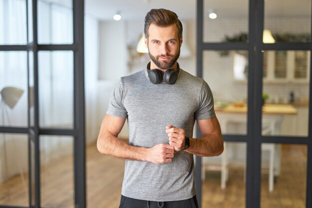 Monsieur barbu avec des écouteurs sans fil autour du cou mettant une montre intelligente sur son poignet et regardant la caméra