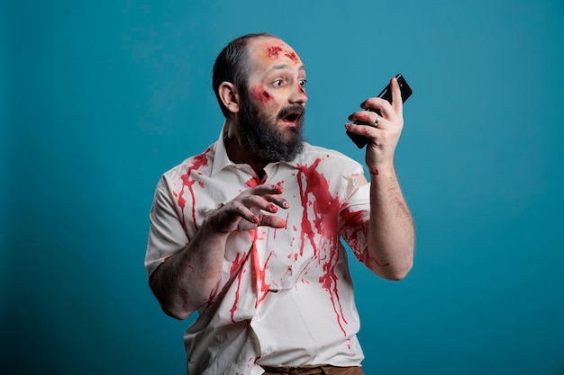 Monstre effrayant d'halloween tenant un smartphone en studio, utilisant une application de téléphone portable tout en étant agressif et effrayant. Zombie mort-vivant effrayant avec des blessures sanglantes, cadavre cruel avec téléphone.