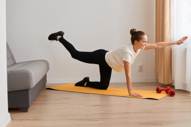 Monter une femme active sportive portant des vêtements de sport debout dans une pose de planche avec la jambe du bras levée, faisant de l'entraînement de yoga, étirant l'exercice de base sur un tapis au sol dans le salon à la maison.
