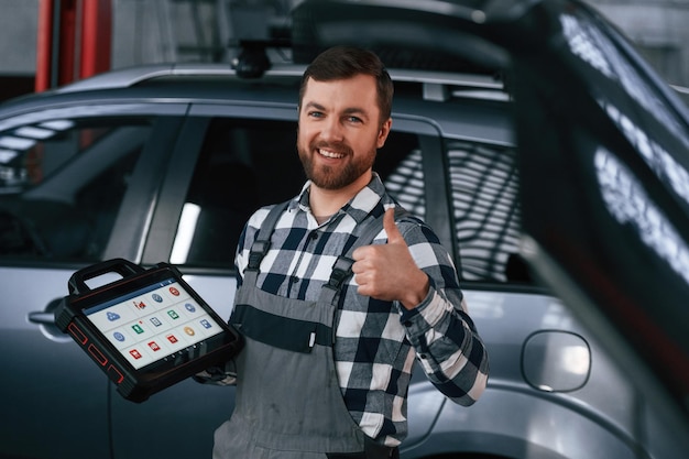 Montrant le geste et souriant Tenant la tablette L'homme en uniforme travaille dans le salon de l'automobile