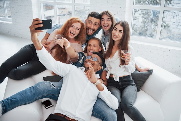 Montrer les langues. Joyeux jeunes amis s'amusant et buvant à l'intérieur blanc