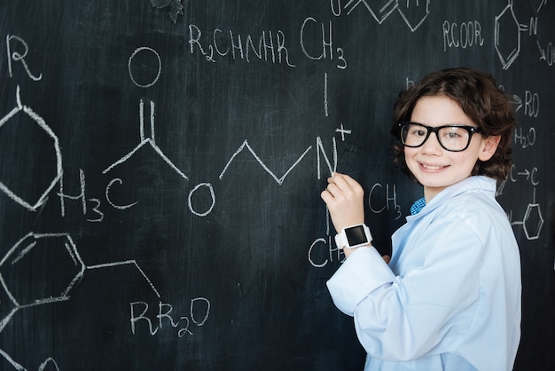 Montrer mes capacités. Heureux élève doué intelligent debout dans le laboratoire tout en profitant d'un cours de chimie et en prenant des notes sur le tableau noir