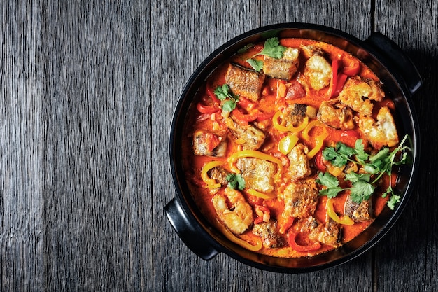 Moqueca baiana - ragoût de poisson brésilien de poisson blanc au poivron doux, citron vert, tomates hachées, lait de coco, servi dans un plat noir avec de la coriandre fraîche sur une table en bois sombre, vue de dessus, espace pour copie