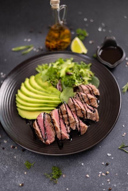 Photo un morceau de filet de thon cuit au gril avec de l'avocat tranché et de la salade sur une plaque de céramique noire