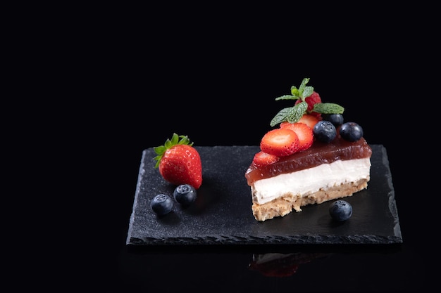 Morceau de gâteau au fromage avec des fraises fraîches et des bleuets isolés servis sur une plaque en ardoise sur fond noir avec espace pour copie. Concept de boulangerie maison.