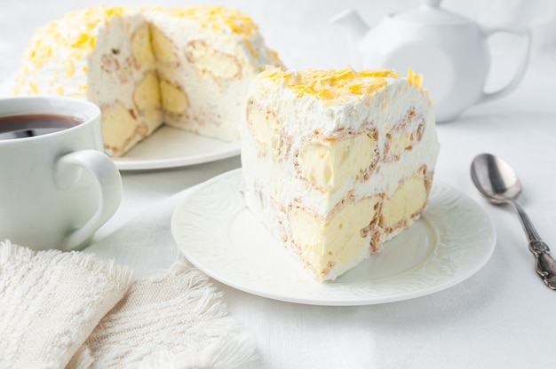 Un morceau de gâteau à la crème fouettée Décoré de pépites de chocolat jaune Fait maison Sur une assiette blanche Près d'une cuillère et d'une tasse Au fond se trouve une assiette avec un gâteau et une théière blanche