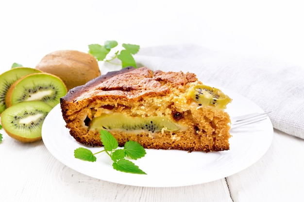 Un morceau de gâteau sucré avec du kiwi et du miel, de la menthe et une fourchette dans une assiette, une serviette sur un fond de planche de bois