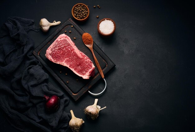 Morceau juteux cru de viande de boeuf sur l'os se trouve sur une planche à découper en bois épices pour la cuisson sur fond noir Filet de viande espace de copie de New York