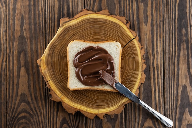 Photo un morceau de pain grillé tartiné de chocolat noisette tartiné avec un couteau sur une vue de dessus de table en bois