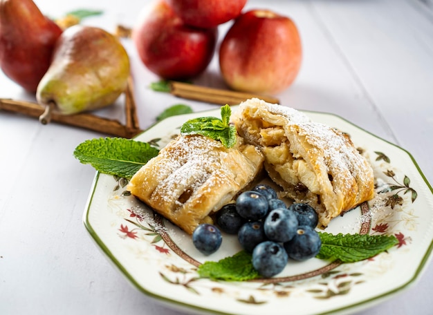 Un morceau traditionnel de strudel aux pommes avec du sucre en poudre menthe myrtille et cannelle au premier plan sur une table