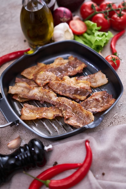 Morceaux de bacon frits dans une poêle et légumes sur une table en pierre ou en béton de cuisine domestique