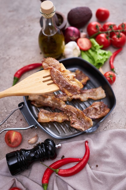 Morceaux de bacon frits dans une poêle et légumes sur une table en pierre ou en béton de cuisine domestique
