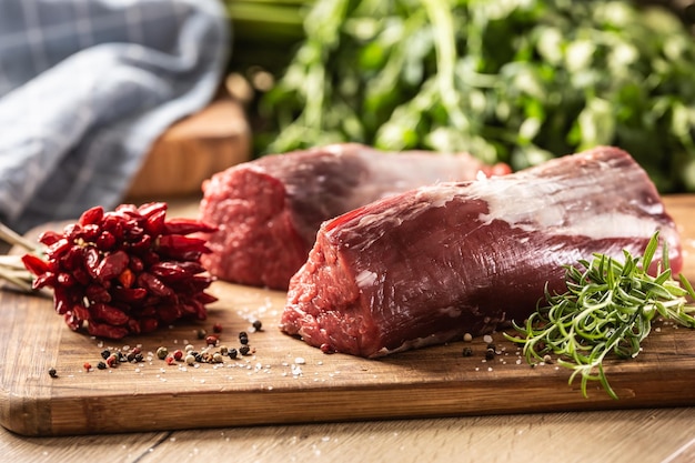 Morceaux crus de viande de surlonge rouge sur une planche de bois à côté de romarin frais et de piments