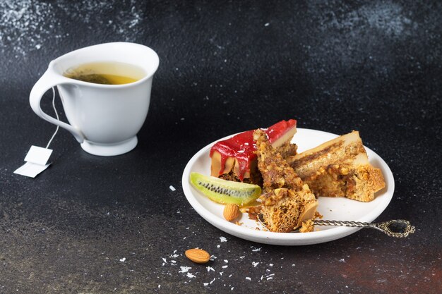 Morceaux de gâteau cassés avec glaçage à la fraise, copeaux de noix de coco et amandes sur une assiette blanche et une tasse de thé vert