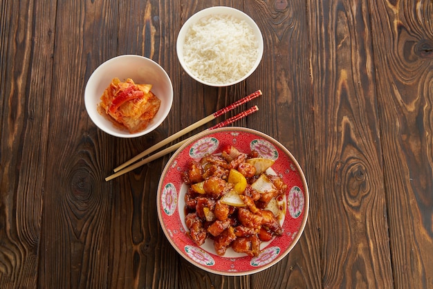 Morceaux de porc frit aigre-doux avec poivrons, oignons et ananas sur plaque rouge avec des baguettes, sur la vieille table en bois avec du riz dans un bol blanc, et soupe traditionnelle, concept de cuisine asiatique