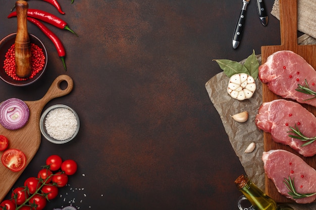 Morceaux de steak de porc cru sur une planche à découper avec tomates cerises, romarin, ail, poivron rouge, feuille de laurier, oignon, mortier de sel et d&#39;épices sur fond brun rouillé