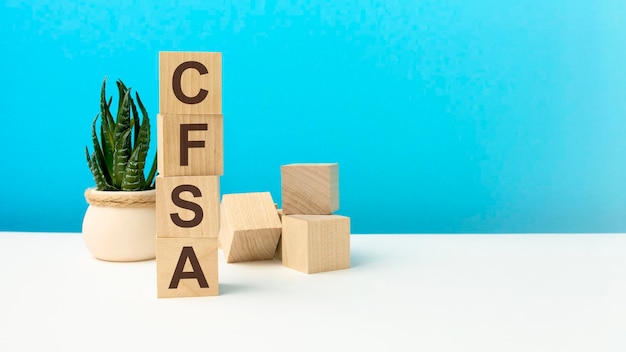 Le mot CFSA est écrit sur des cubes en bois closeup fond bleu vif table de surface blanche en arrière-plan est un cube en bois et une fleur verte Vérificateur des services financiers certifié CFSA