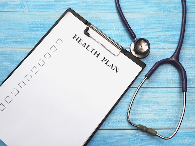 Mot de plan de santé sur le presse-papiers avec du papier blanc et un stéthoscope sur fond de bois bleu. Concept de soins de santé et médical.
