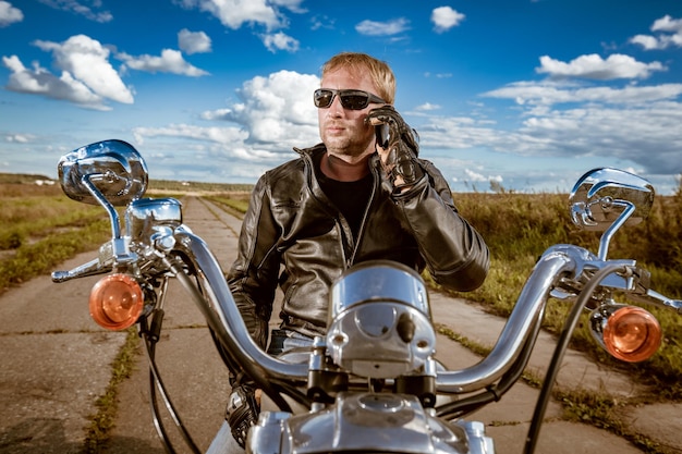 Motard parlant sur un smartphone. Homme motard portant une veste en cuir et des lunettes de soleil assis sur sa moto.