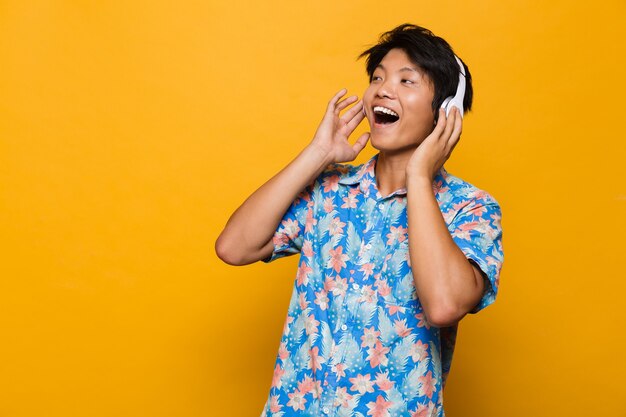 Émotionnel excité jeune homme asiatique debout isolé sur un espace jaune, écouter de la musique avec des écouteurs.