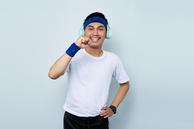 Émotionnel jeune homme asiatique instructeur de conditionnement physique sportif en bandeau bleu et t-shirt blanc avec un casque faire le geste gagnant serrer le poing célébrant isolé sur fond blanc