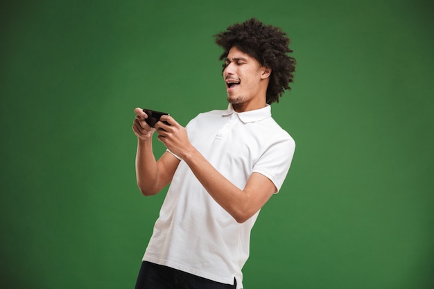 Émotionnel jeune homme bouclé africain jouer à des jeux par téléphone mobile.