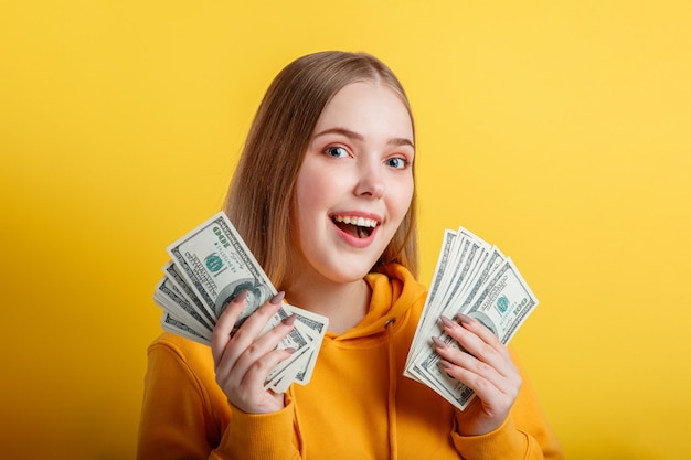 Émotionnelle heureuse adolescente blonde gagner de l'argent en espèces en tenant des dollars dans les mains isolés sur fond jaune de couleur. Portrait jeune femme excitée avec fan de billets d'argent.