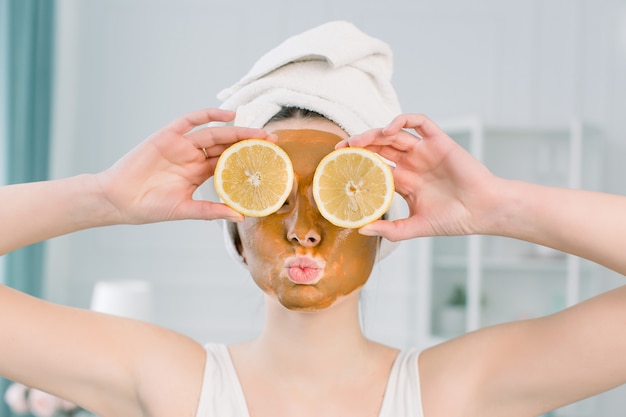 Émotionnelle jeune femme en serviette blanche sur la tête et avec un masque facial et des moitiés de citron mûr sur un espace blanc. Photo d'une femme recevant des soins spa. Concept de beauté et de soins de la peau