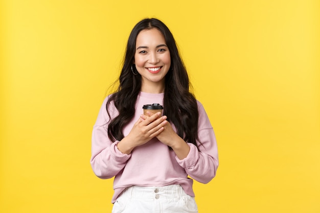 Émotions, concept de loisirs et de beauté. Belle femme asiatique avec une tasse de café à emporter profitant des week-ends d'été, souriant ravi et mignon, boire des boissons sur le mur jaune