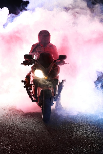 Photo motocycliste dans un costume de veste en cuir à travers la fumée rouge et bleue sur une moto la nuit