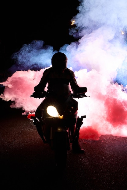 Photo motocycliste dans un costume de veste en cuir à travers la fumée rouge et bleue sur une moto la nuit