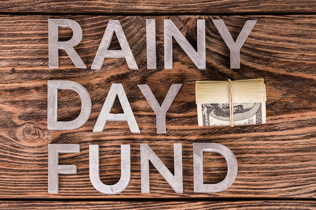 Mots RAINY DAY FUND posés sur une surface en bois par des lettres métalliques avec un petit rouleau de billets en dollars américains à plat au-dessus de la vue