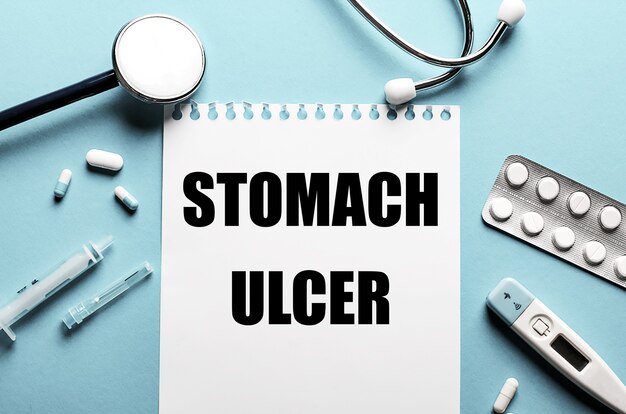 Les mots STOMACH ULCER écrits sur un bloc-notes blanc sur une surface bleue près d'un stéthoscope, d'une seringue, d'un thermomètre électronique et de pilules. Concept médical