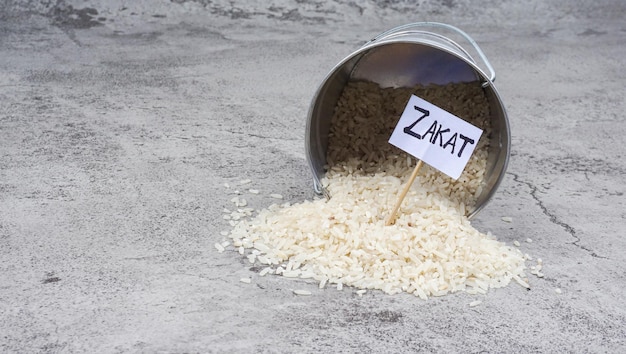 Mots Zakat avec tas de riz blanc dans un seau en métal contre fond texturé ciment