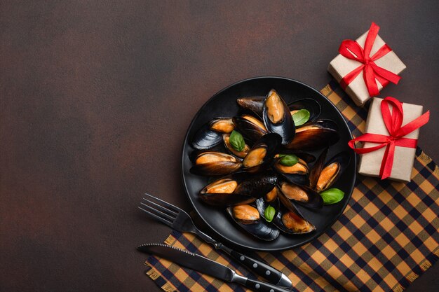 Moules de fruits de mer et feuilles de basilic dans une assiette noire avec des boîtes de fourchette, couteau et cadeau sur une serviette et fond rouillé